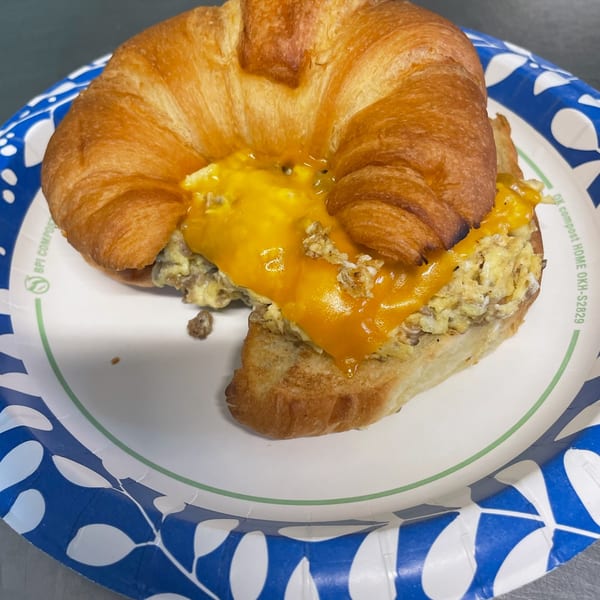 Croissant breakfast sandwich with scrambled eggs, melted cheese, and sausage on a paper plate.