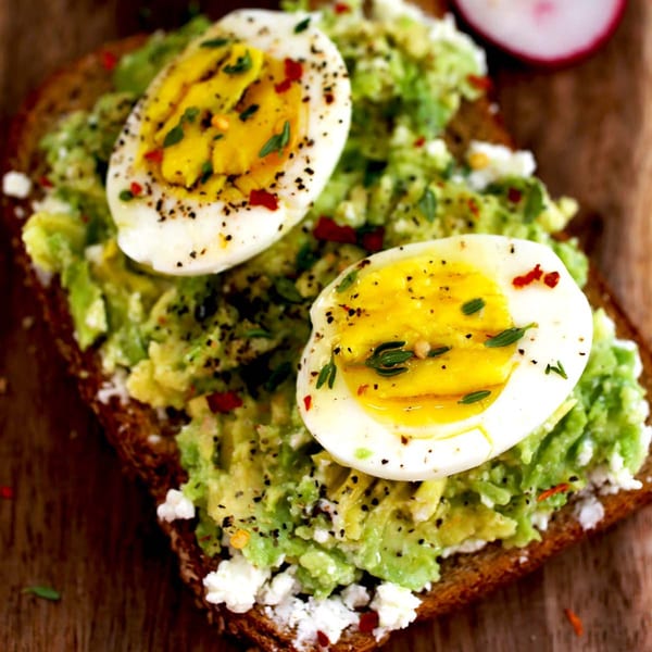Avocado toast with two halved hard-boiled eggs, crumbled cheese, and spices on a wooden board.